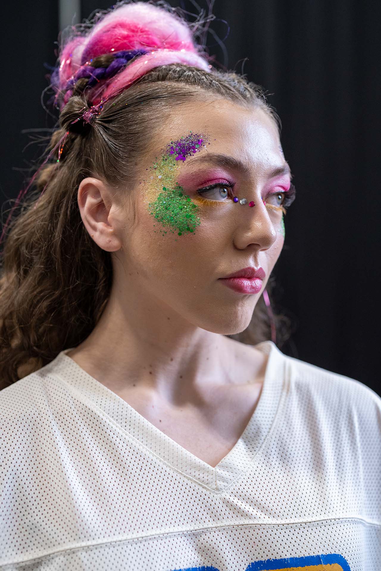 Backstage portrait of a model with pink and green glitter eye makeup in a white top
