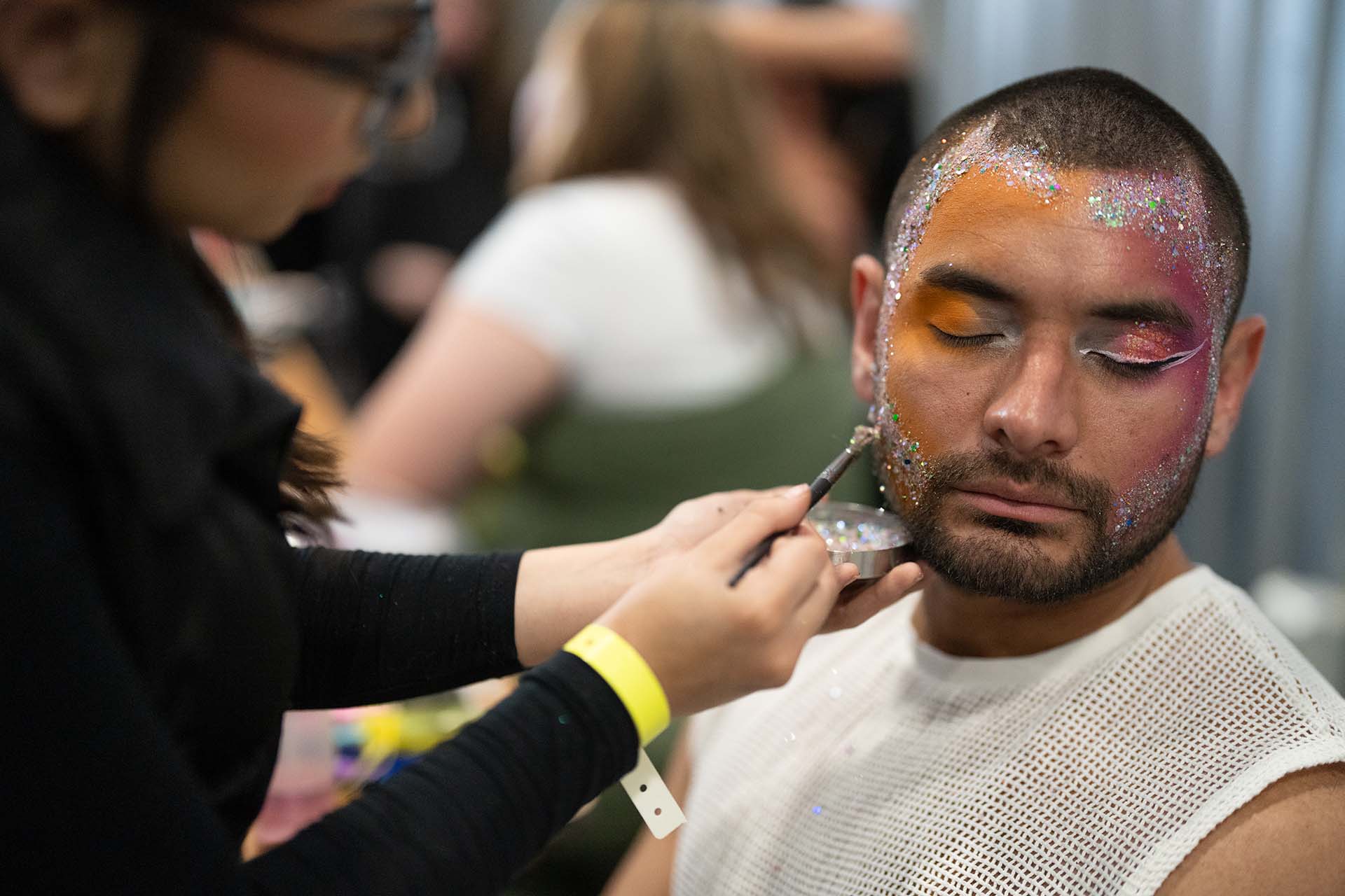 Backstage photo of a makeup artist applying colorful eye makeup to a male model