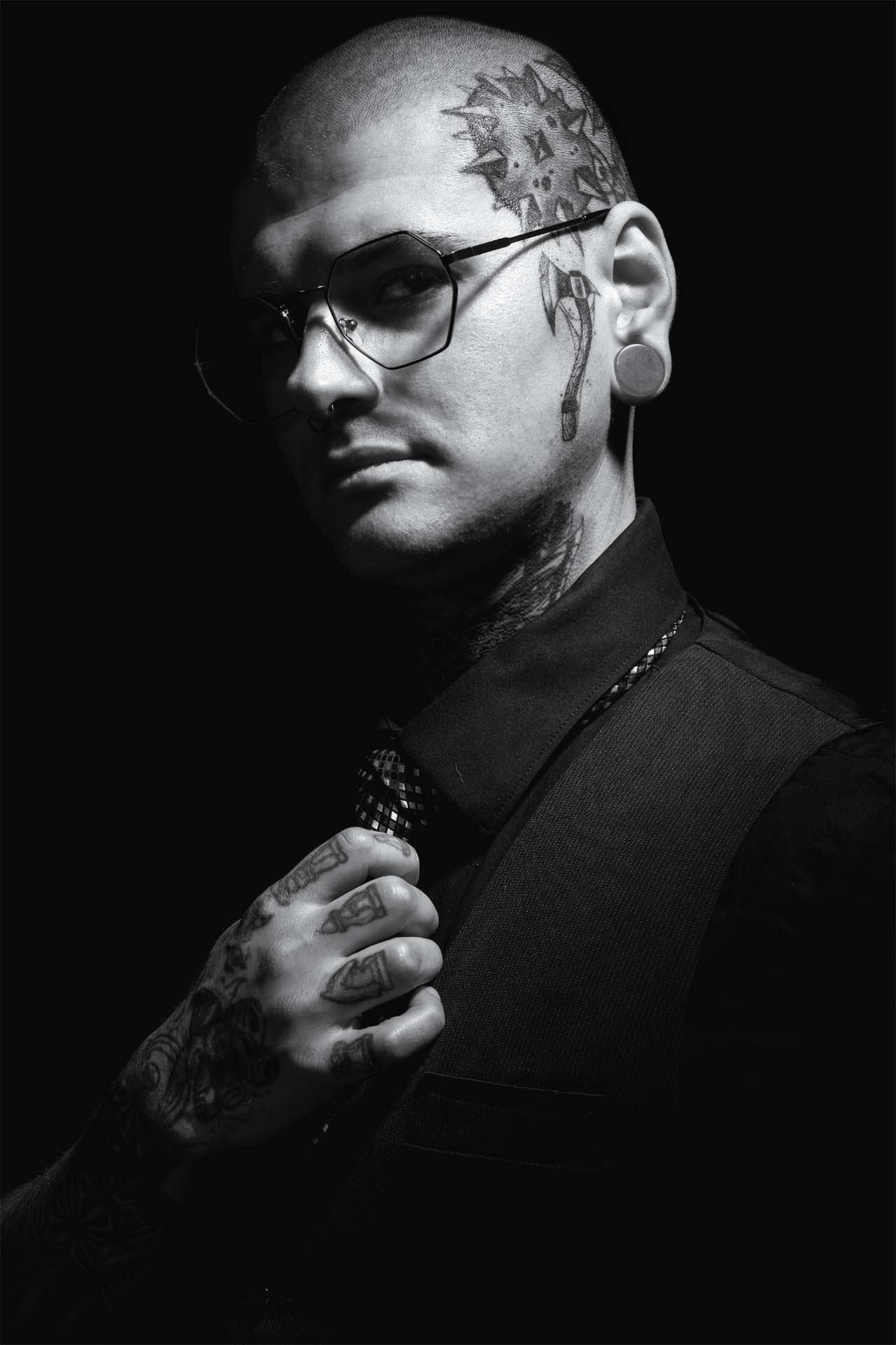 Black-and-white portrait of a tattoo artist in glasses with hand near his face, dramatic studio lighting