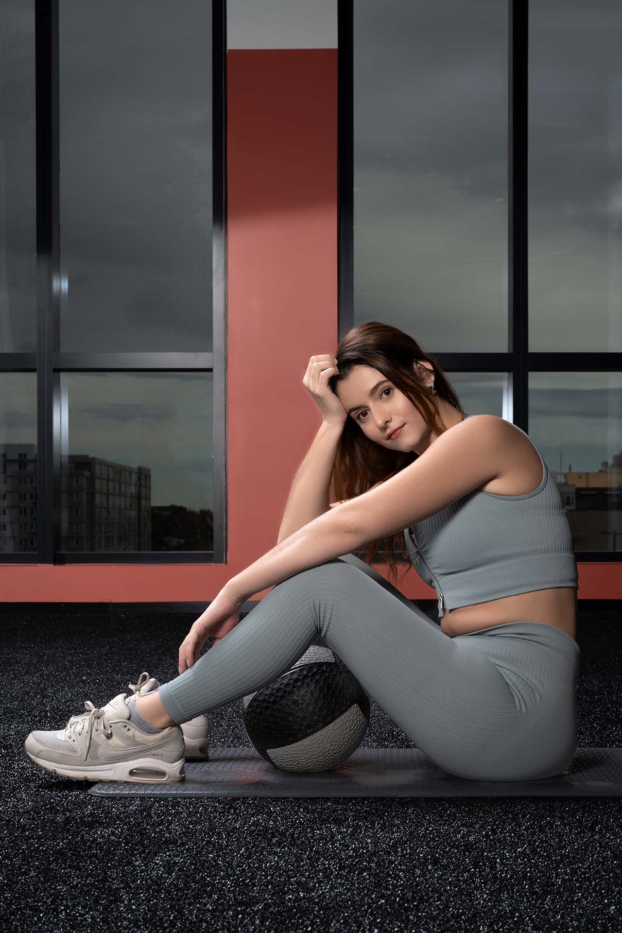 Woman in grey athletic wear seated on a yoga mat, seated with a medicine ball, city window backdrop