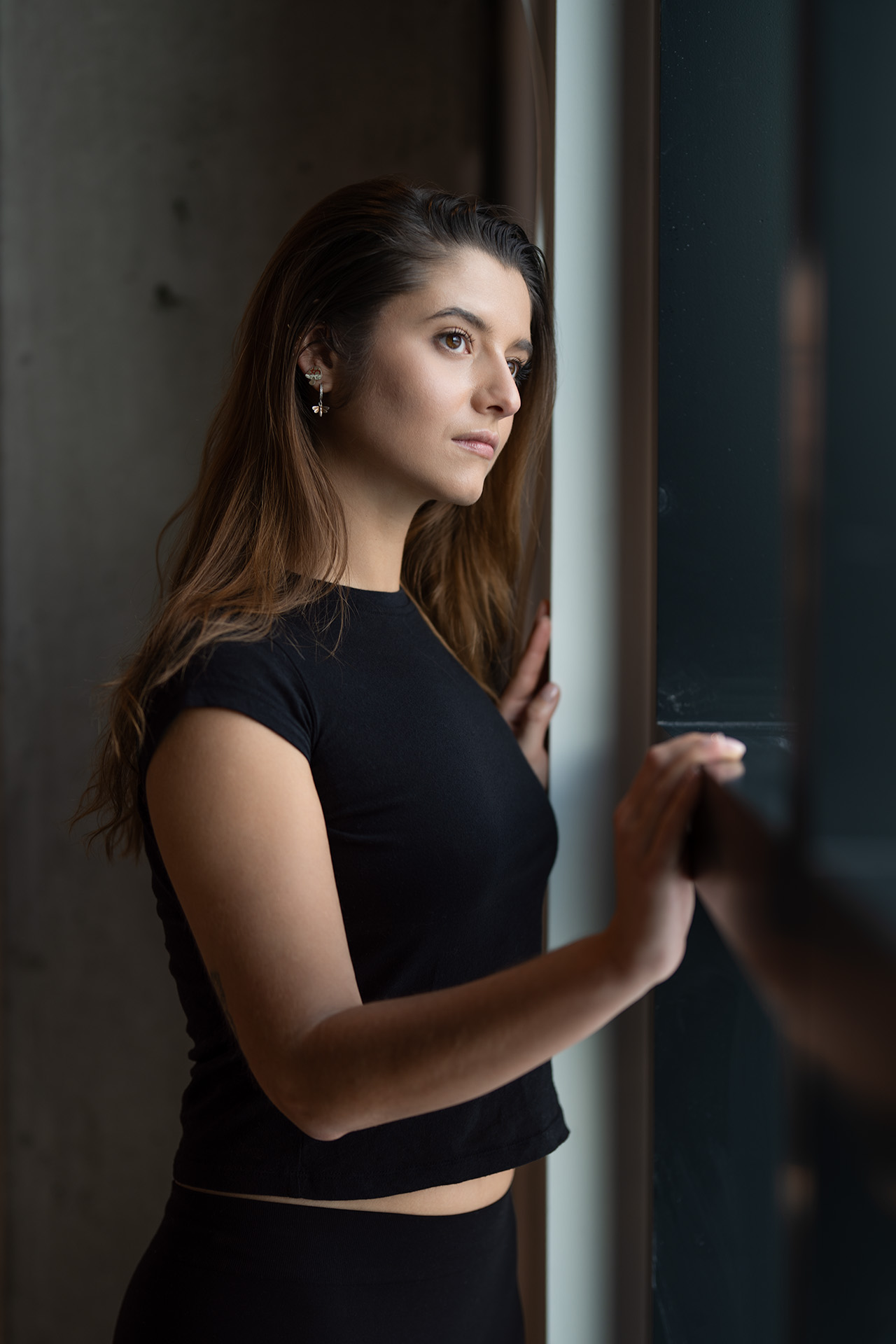 Side-profile portrait of a model in a black top leaning against a window frame, moody lighting