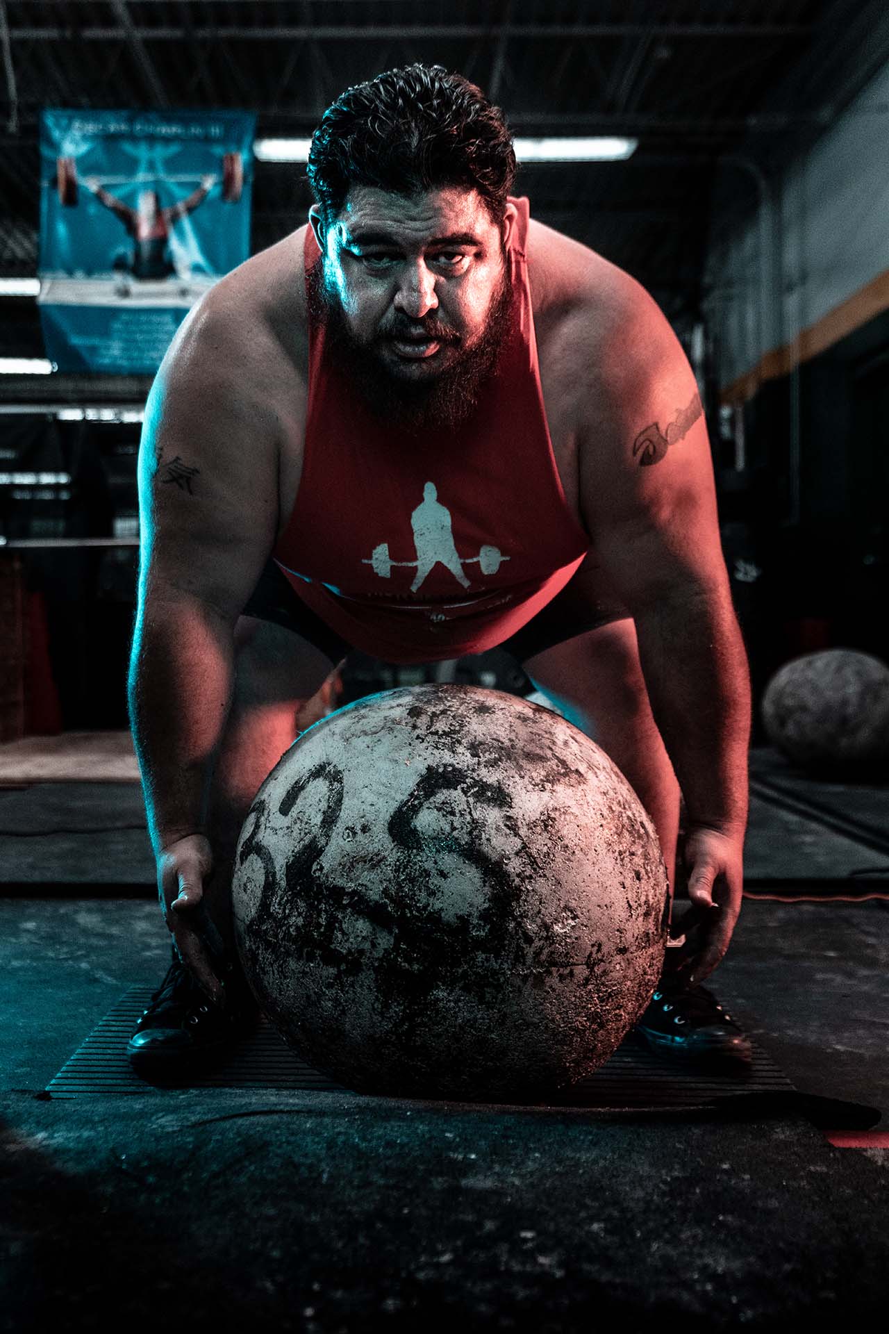Strongman athlete in a red tank top lifting an atlas stone in an industrial warehouse