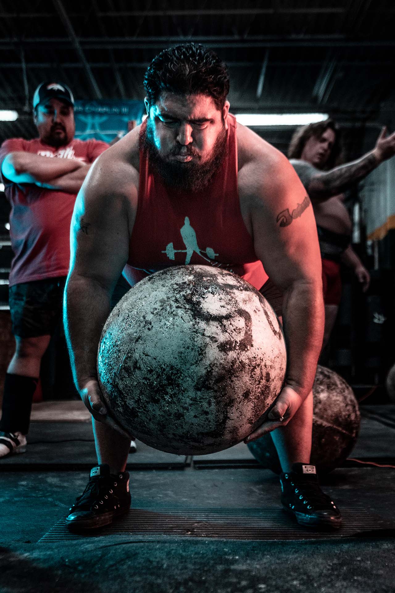 Strongman athlete lifting an atlas stone with spotters visible in the background, industrial gym