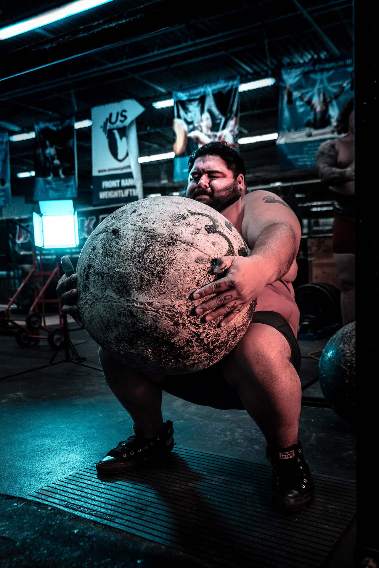 Strongman athlete lifting an atlas stone under dramatic teal lighting, industrial gym