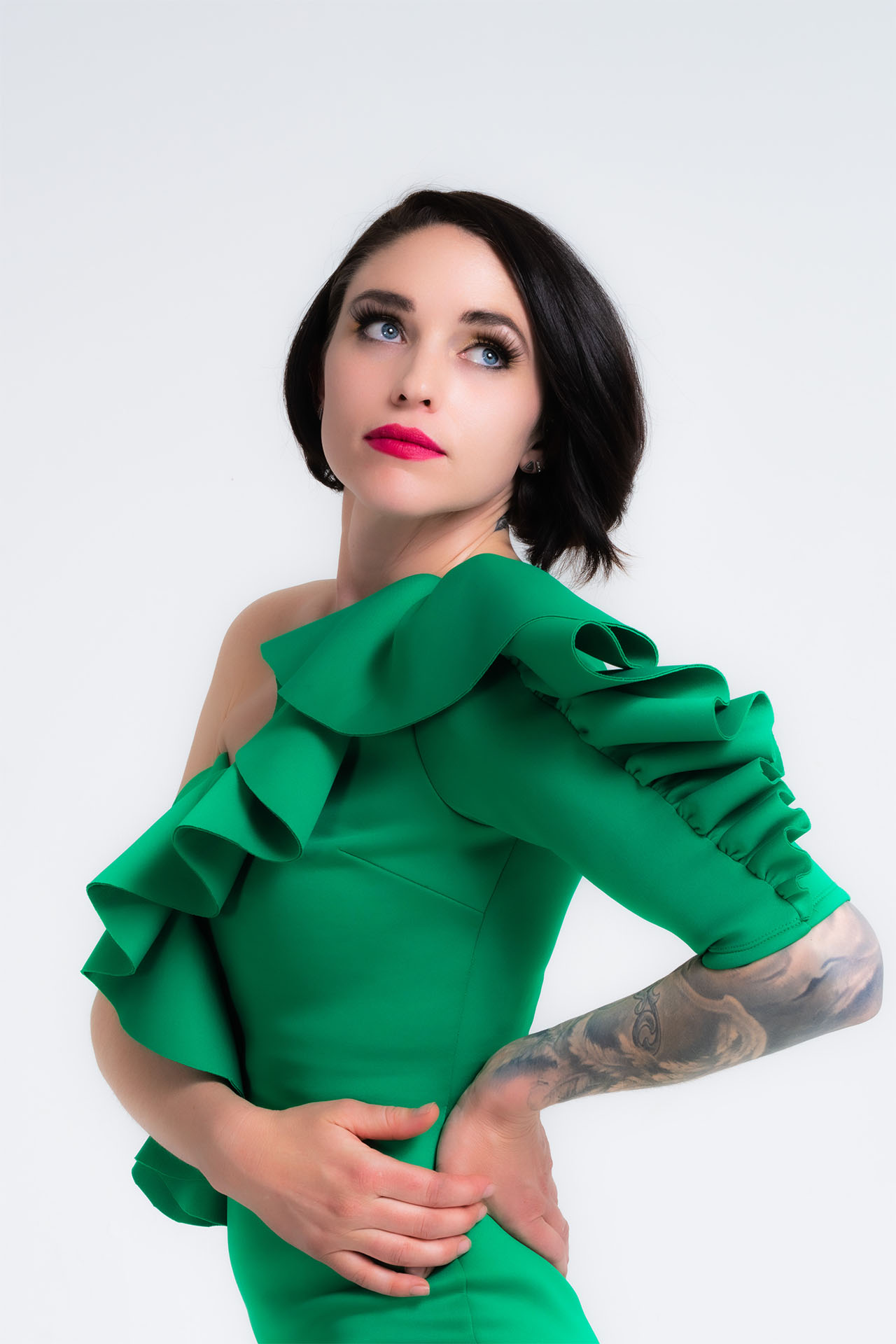 Model in a green ruffled off-shoulder dress with bold makeup and visible tattoos, studio portrait