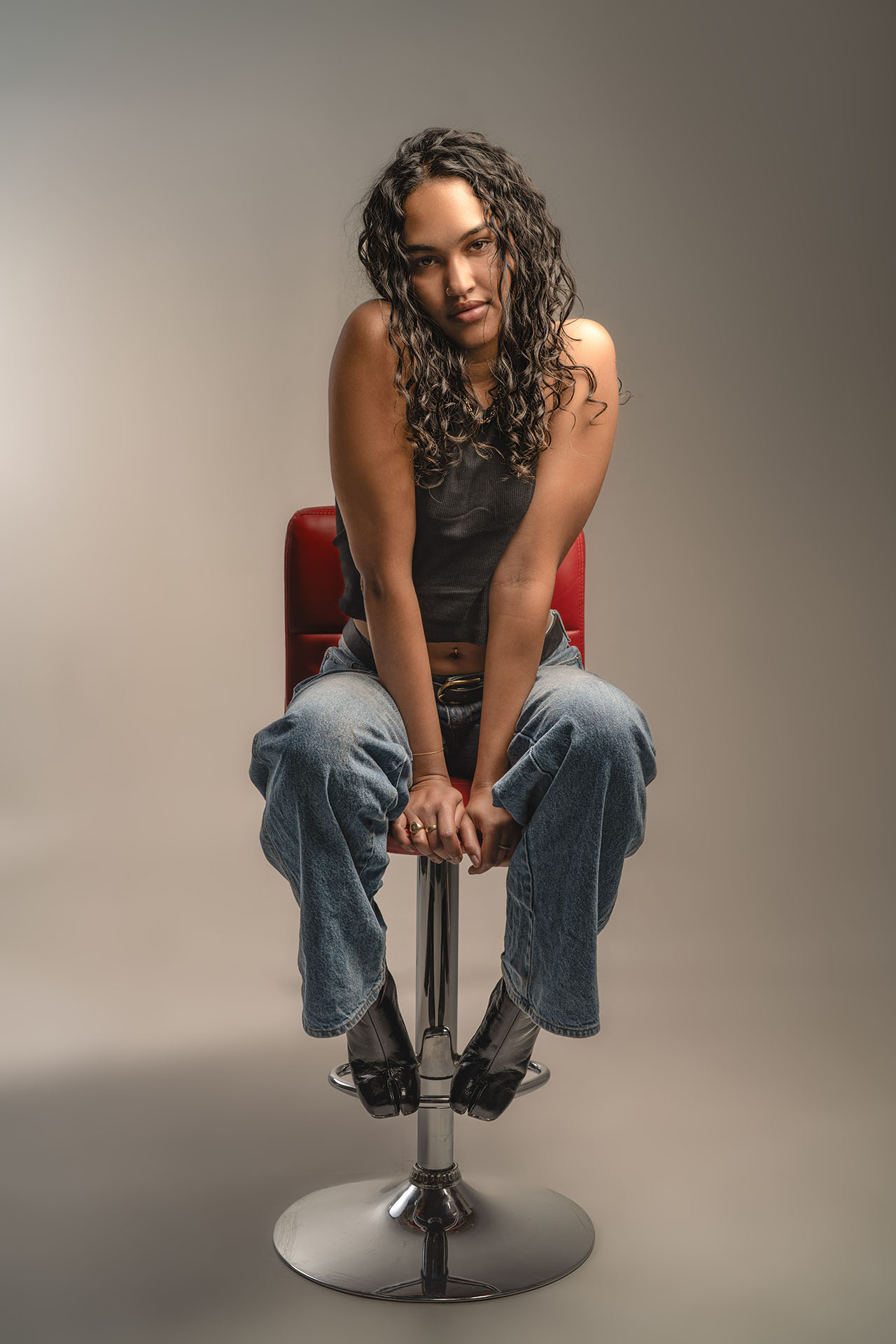 Model seated on a stool in jeans and a black top, gray studio backdrop
