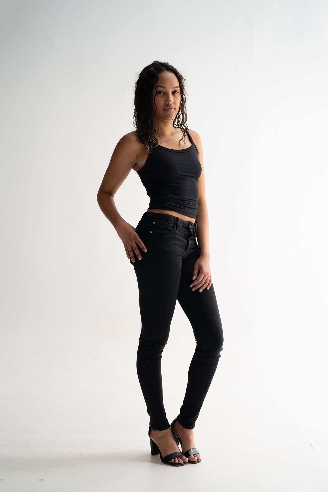 Model in a black tank top and jeans posing for portfolio digitals against a white studio background