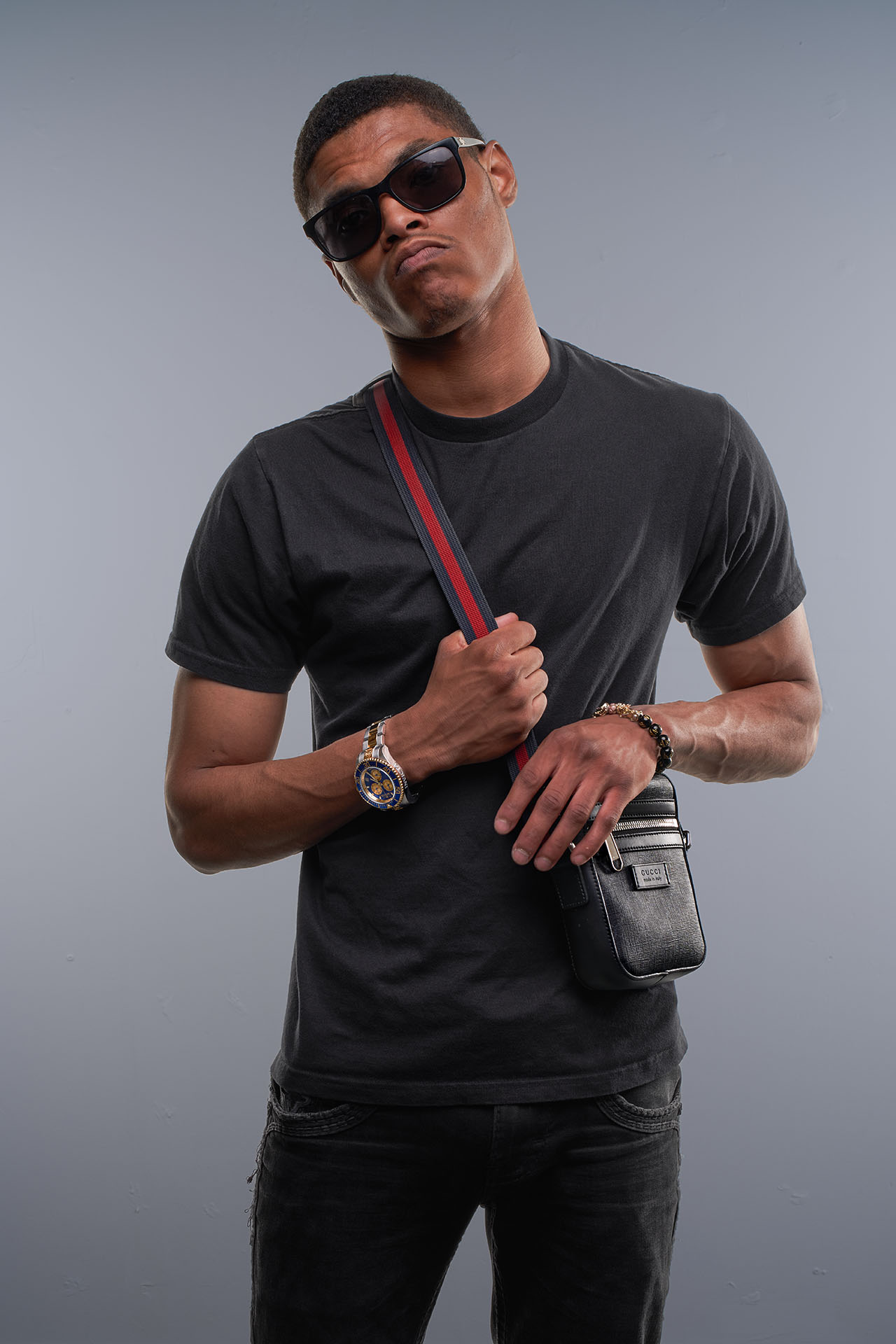 Male model in sunglasses and a black t-shirt with a red crossbody strap bag, gray studio background