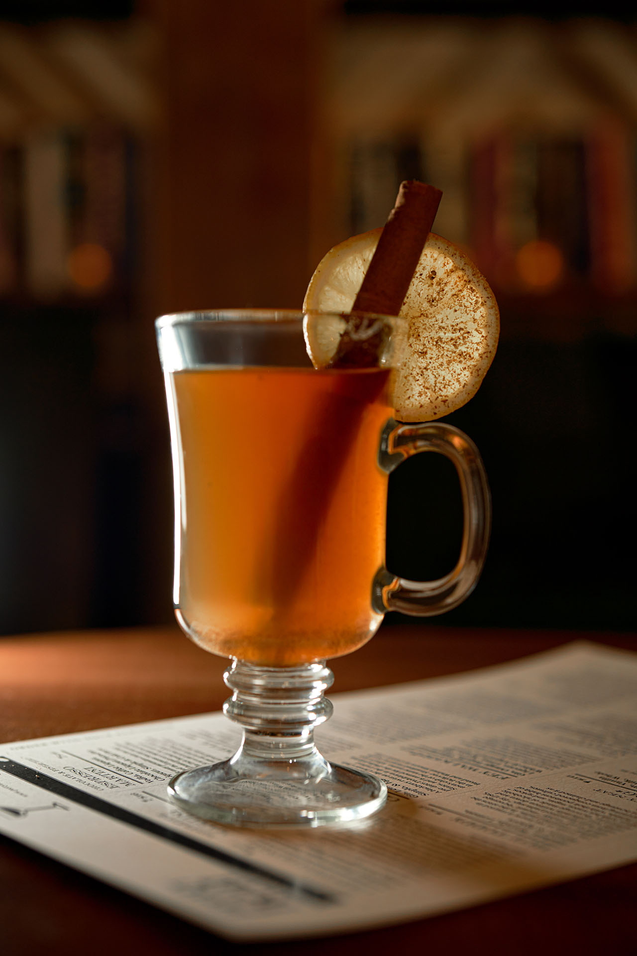 Hot toddy in a glass mug with a cinnamon stick garnish, first styling pass on a newspaper prop