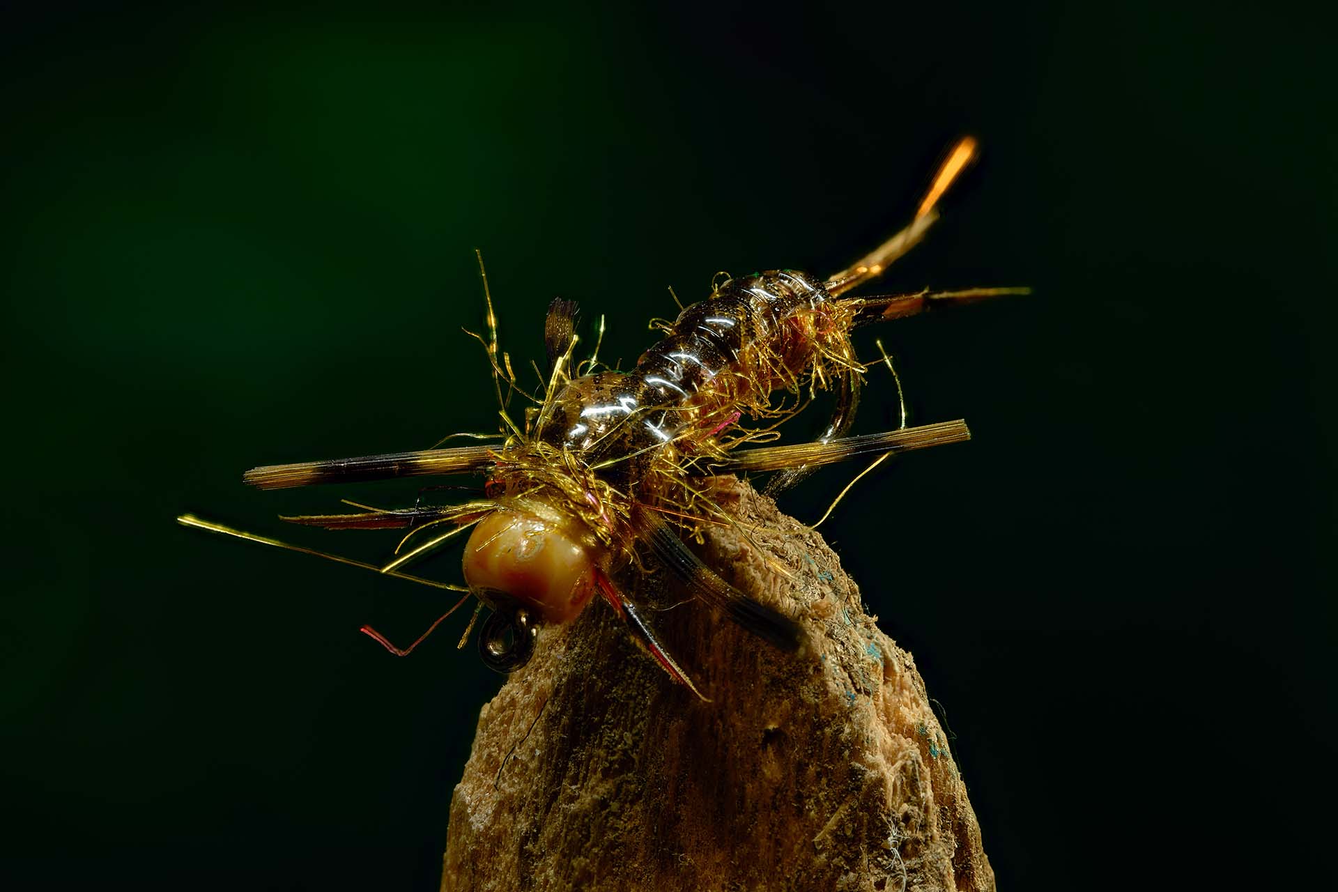 Macro product photo of a tan hand-tied fishing fly on a wood perch against a dark background