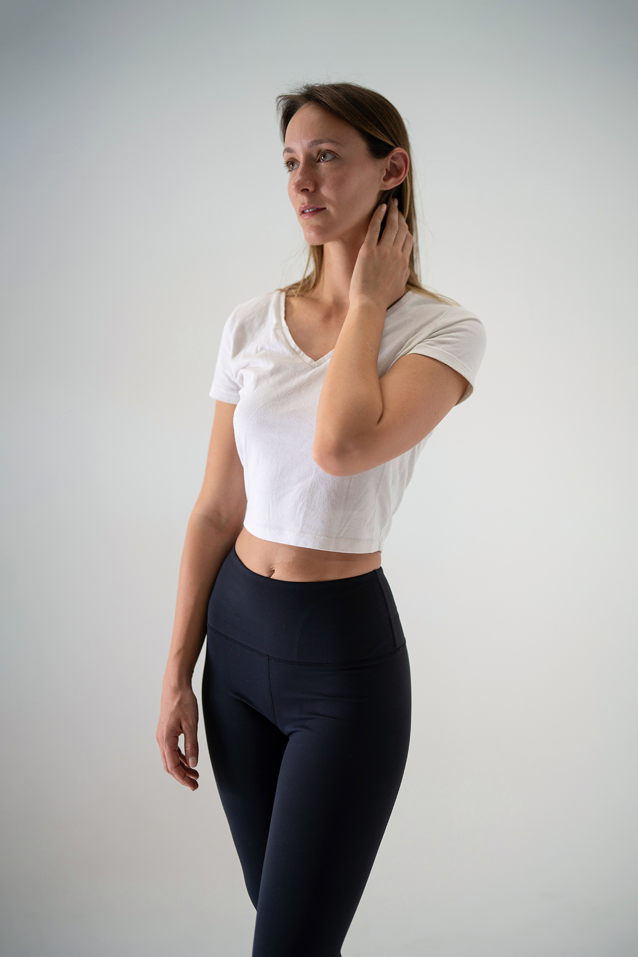 Model portfolio digital, hand raised near the neck, white crop top and black leggings, cropped at mid-thigh