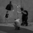 Black and white behind-the-scenes photo of a fashion shoot in a parking garage: a model in a chain-draped black outfit and metallic thigh-high boots stands under a softbox light while a photographer crouches in the foreground and two crew members adjust an overhead boom light.