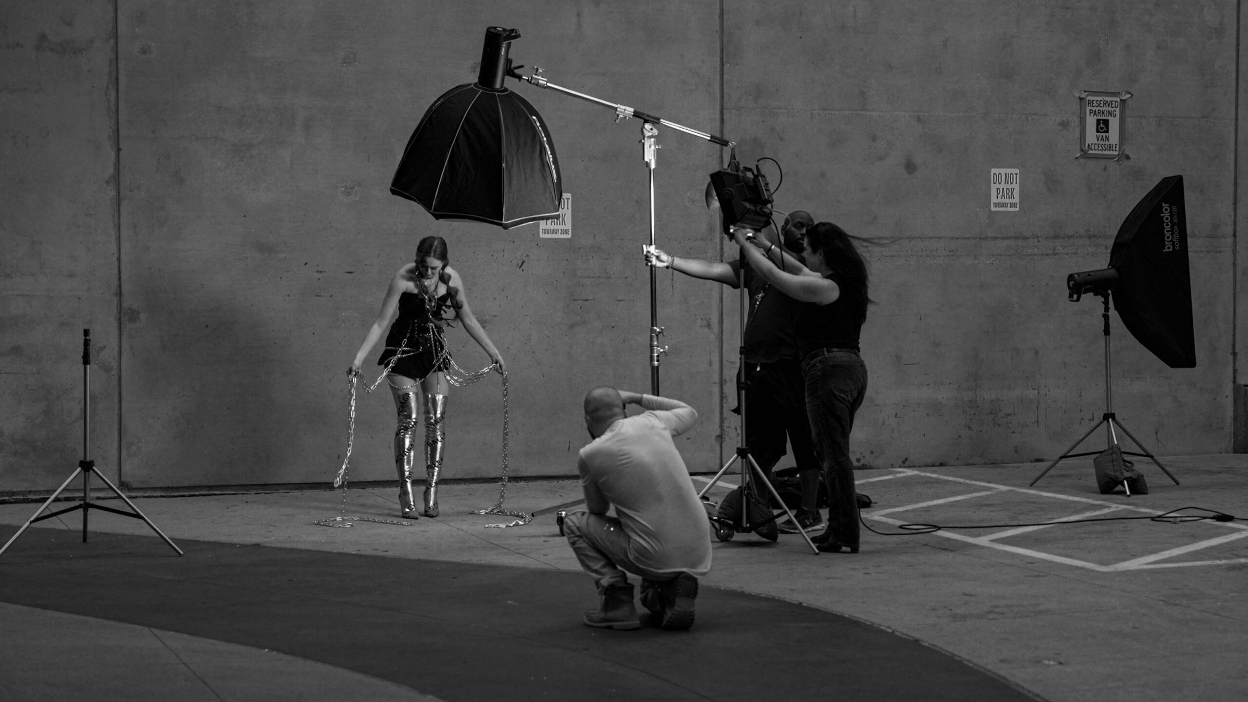 Black and white behind-the-scenes photo of a fashion shoot in a parking garage: a model in a chain-draped black outfit and metallic thigh-high boots stands under a softbox light while a photographer crouches in the foreground and two crew members adjust an overhead boom light.